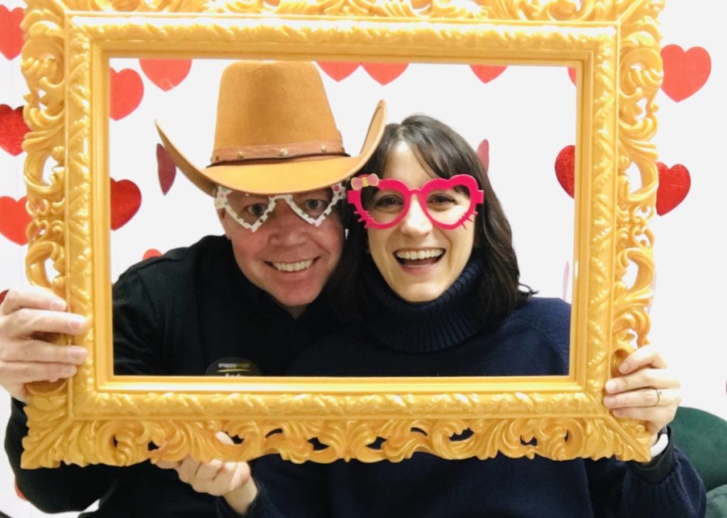 A man and a woman pose with a cut out photo frame, wearing silly glasses.