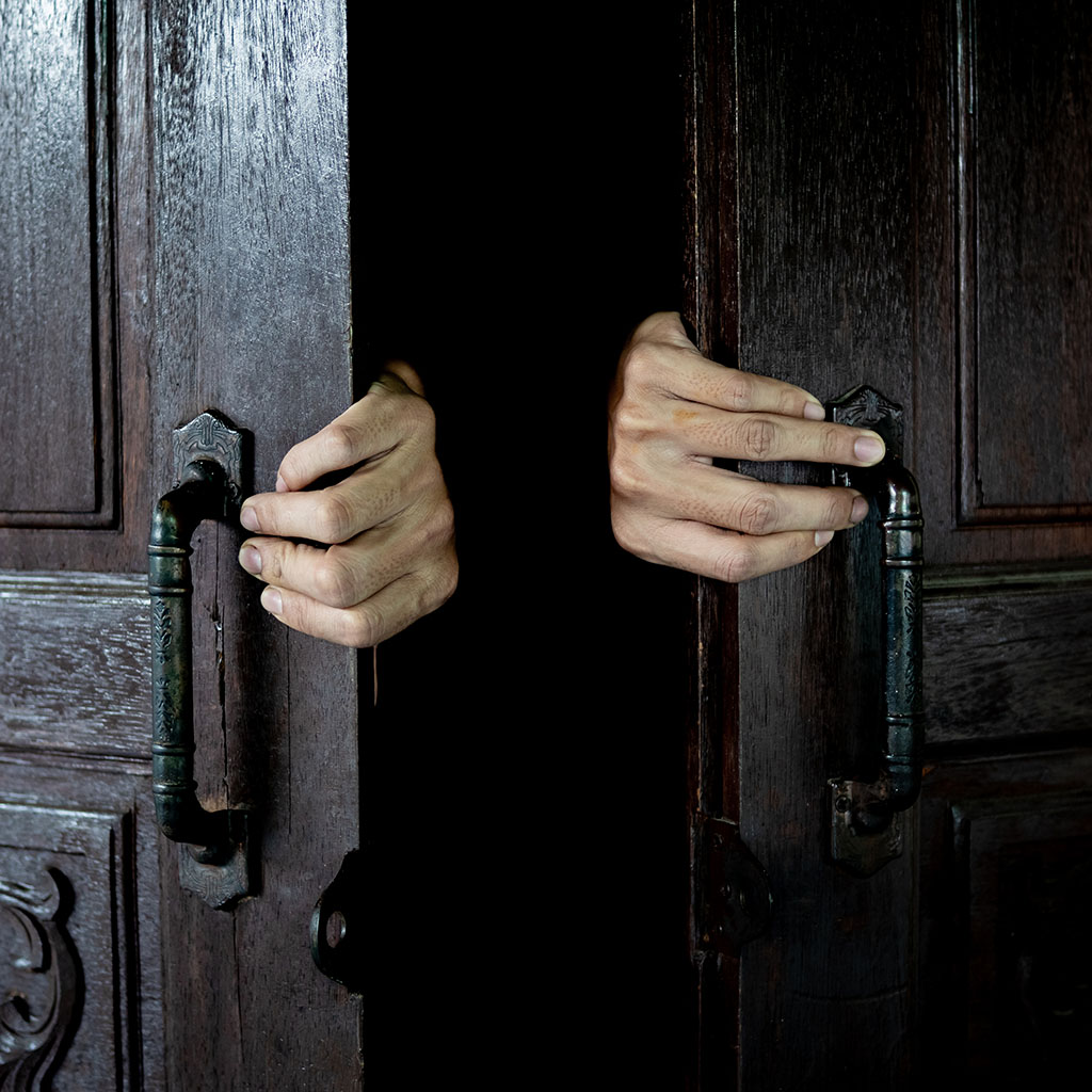 Hands open the wooden door from the inside of the dark room.