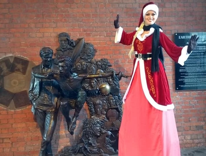 lady wearing red santa cloak on stilts next to Bowie statue